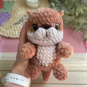 May include: A small, plush, crocheted otter toy made from soft, light brown yarn. It features a white belly and muzzle, a dark brown nose, and large, round, black plastic eyes with gold glitter accents. The toy has a textured, fuzzy appearance.