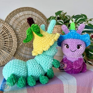 Puede incluir: Dos peluches de oruga hechos a mano con ganchillo. Uno es turquesa con un sombrero de flores amarillo y detalles verdes. El otro es morado con un sombrero azul y detalles rosas. Los juguetes est&aacute;n sobre una superficie a rayas rosas y blancas.