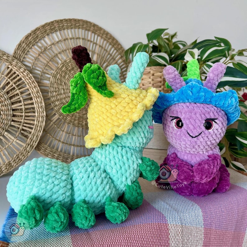 Puede incluir: Dos peluches de oruga hechos a mano con ganchillo. Uno es turquesa con un sombrero de flores amarillo y detalles verdes. El otro es morado con un sombrero azul y detalles rosas. Los juguetes est&aacute;n sobre una superficie a rayas rosas y blancas.