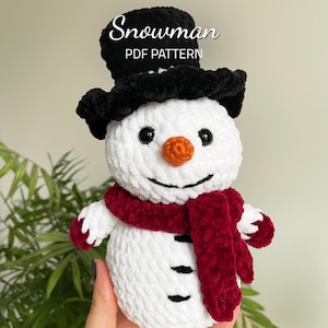 Puede incluir: Un muñeco de nieve de crochet blanco con un sombrero de copa negro, una bufanda roja y ojos y boca negros. El muñeco de nieve tiene una nariz naranja y botones negros. El texto "Snowman PDF Pattern" está por encima del muñeco de nieve.