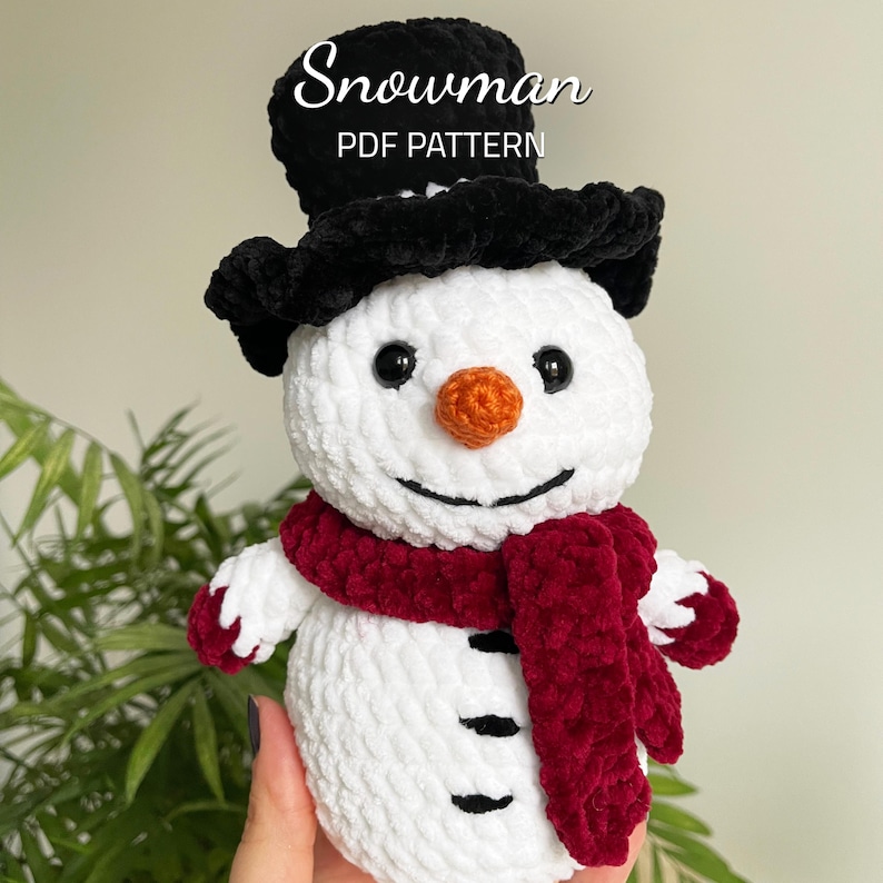 May include: A white crocheted snowman with a black top hat, red scarf, and black eyes and mouth. The snowman has an orange nose and black buttons. The text "Snowman PDF Pattern" is above the snowman.