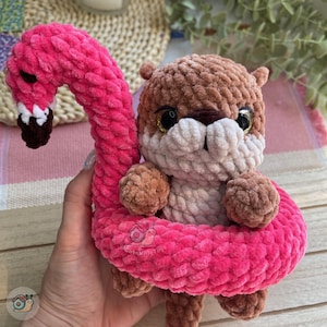 May include: A crochet plush toy featuring a brown otter with a cream-colored muzzle and large black eyes, nestled inside a bright pink crochet flamingo floatie. The flamingo has a long, curved neck, a black beak with white accents, and a soft texture.