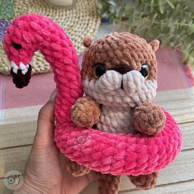 May include: A crochet plush toy featuring a brown otter with a cream-colored muzzle and large black eyes, nestled inside a bright pink crochet flamingo floatie. The flamingo has a long, curved neck, a black beak with white accents, and a soft texture.