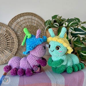 Puede incluir: Dos peluches de oruga hechos a mano con ganchillo. Uno es morado con un sombrero azul y el otro es verde azulado con un sombrero amarillo. Ambos tienen caras bordadas y est&aacute;n sobre una superficie rayada. Los juguetes son de @CuteVilleToys.