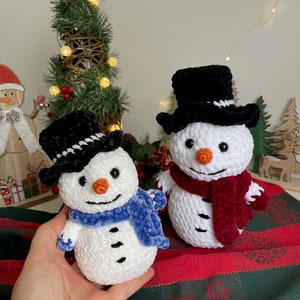 May include: Two crocheted snowmen with black top hats and scarves. One snowman has a blue scarf and the other has a burgundy scarf. The snowmen are sitting on a red and green striped fabric.