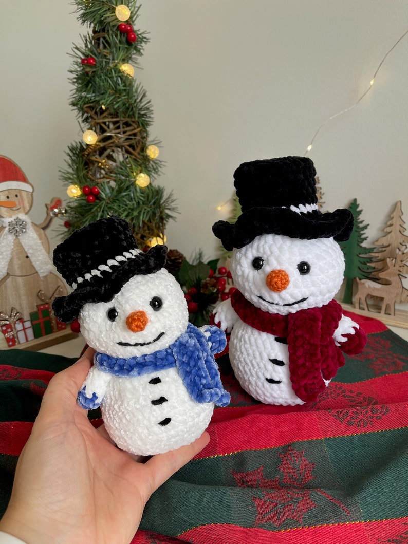 May include: Two crocheted snowmen with black top hats and scarves. One snowman has a blue scarf and the other has a burgundy scarf. The snowmen are sitting on a red and green striped fabric.