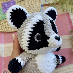 May include: A plush raccoon toy made of soft, chunky yarn in shades of gray, black, and white. The stuffed animal features large, dark, glossy eyes with silver glitter accents, a black plastic nose, and a white belly patch with the text CuteVilleToys.