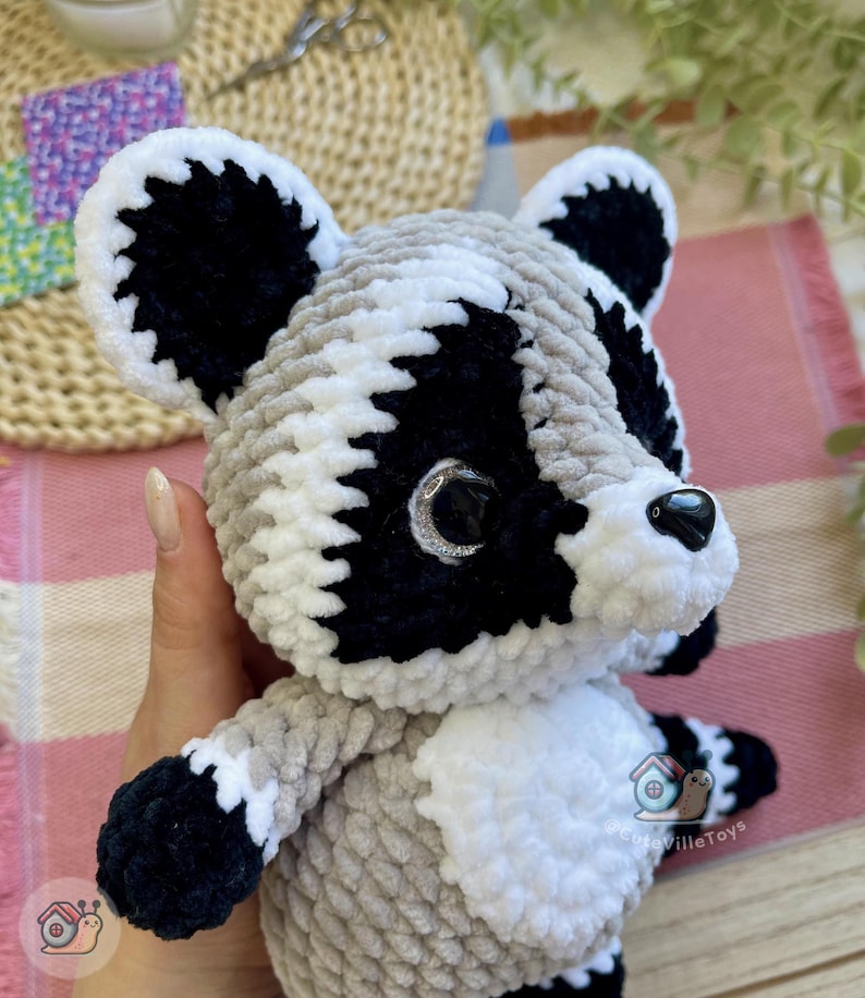 May include: A plush raccoon toy made of soft, chunky yarn in shades of gray, black, and white. The stuffed animal features large, dark, glossy eyes with silver glitter accents, a black plastic nose, and a white belly patch with the text CuteVilleToys.