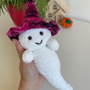 Op de afbeelding: Witte gehaakte geest knuffel met een roze en paarse fluwelen heksenhoed met zwarte ogen en een glimlachende mond.