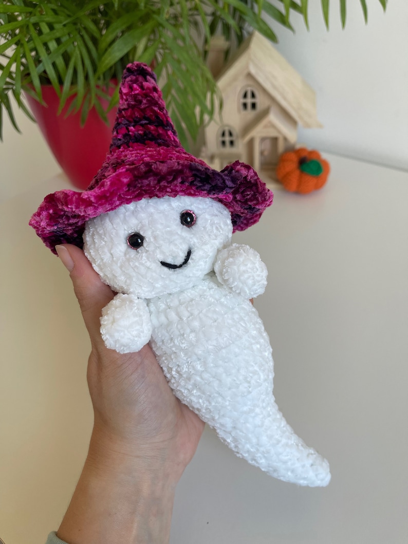 Op de afbeelding: Witte gehaakte geest knuffel met een roze en paarse fluwelen heksenhoed met zwarte ogen en een glimlachende mond.