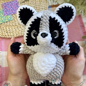 May include: A plush raccoon toy made of soft, textured yarn in shades of light gray, white, and black. The raccoon features large, glossy black eyes with silver accents, a small black nose, and a white belly patch. The toy has black paws and ears.