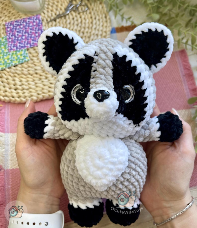 May include: A plush raccoon toy made of soft, textured yarn in shades of light gray, white, and black. The raccoon features large, glossy black eyes with silver accents, a small black nose, and a white belly patch. The toy has black paws and ears.
