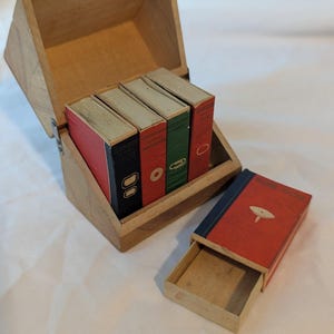 May include: A wooden box with a hinged lid containing four small rectangular boxes with red, green, and black covers featuring graphic icons. A fifth matching red box with a blue spine and a spinning top icon sits open in front of the wooden container.