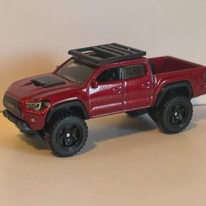 May include: A die-cast, red Toyota Tacoma pickup truck with black wheels and a roof rack. The truck has a detailed design, including a hood scoop and a truck bed. The tires are large and off-road style.