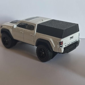 May include: A white toy pickup truck with black wheels and a black bed cover. The truck has the word "Tacoma" printed on the back. The toy truck is a miniature replica of a real vehicle.