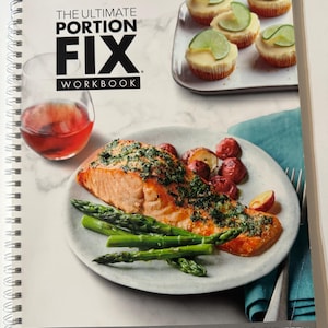 May include: A spiral-bound workbook titled "THE ULTIMATE PORTION FIX WORKBOOK" with a plate of food including salmon, asparagus, and potatoes. A glass of red liquid and mini cheesecakes with lime slices are also visible. The image promotes healthy eating.