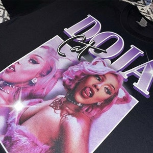 May include: A black t-shirt with a purple and white graphic print of Doja Cat. The graphic features a close-up of Doja Cat's face with pink hair and a cat ear headband. The text "Doja Cat" is printed in a bold, white font.