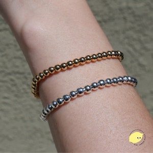 Puede incluir: Dos pulseras de cuentas, una dorada y otra plateada, en la muñeca de una persona. Las pulseras están hechas de pequeñas cuentas redondas.