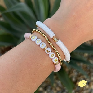 May include: A set of three bracelets, one with white beads and a gold heart charm, one with gold beads and a pink band, and one with white beads spelling out "BOY MOM".