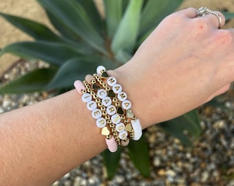 May include: A set of five bracelets with white beads and gold lettering spelling out the names "Mama", "Carson", "Tyler", "Son", and "Love". Each bracelet has a small gold heart charm.