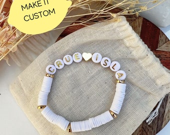 May include: White beaded bracelet with gold letters spelling "TRUE IS LA" and a gold heart charm. The bracelet is on a beige fabric surface.