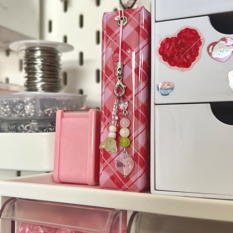 Peut inclure: Un classeur rose et blanc &agrave; carreaux avec un charme perl&eacute; rose et blanc attach&eacute;. Le charme a un cristal en forme de c&oelig;ur en bas et deux petits charmes en forme de fleur au-dessus.
