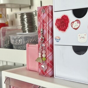 Peut inclure: Un charme en tissu &agrave; carreaux rose et blanc avec un charme argent&eacute; attach&eacute; &agrave; un cordon rose et blanc. Le charme pr&eacute;sente un c&oelig;ur rose avec un cristal transparent au centre et un groupe de perles roses, vertes et blanches.