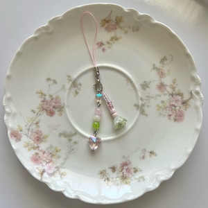 Peut inclure: Un porte-cl&eacute;s de t&eacute;l&eacute;phone en perles roses et vertes avec un cristal en forme de c&oelig;ur et un charme de fleur. Le charme est suspendu &agrave; une ficelle rose sur une assiette blanche &agrave; motif floral.