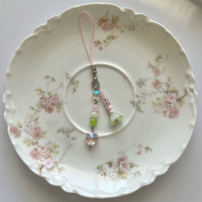 Peut inclure: Un porte-cl&eacute;s de t&eacute;l&eacute;phone en perles roses et vertes avec un cristal en forme de c&oelig;ur et un charme de fleur. Le charme est suspendu &agrave; une ficelle rose sur une assiette blanche &agrave; motif floral.