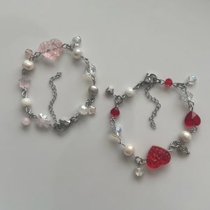 May include: Two delicate bracelets with silver chains and charms. One bracelet features pink heart-shaped beads and the other features red heart-shaped beads.