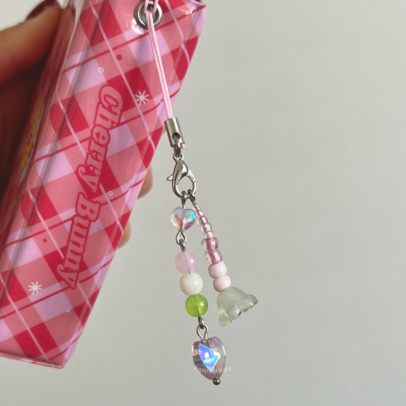 Peut inclure: Un carnet &agrave; carreaux rose et blanc avec un charme en perles rose et blanc attach&eacute;. Le charme comprend un cristal en forme de c&oelig;ur, des perles rose et blanc et deux petits charmes en forme de fleur. Le carnet porte l'inscription "Cherry Bunny" sur la couverture.