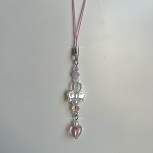 May include: A pink and white phone charm with a silver bow and heart charm. The charm is attached to a pink string.