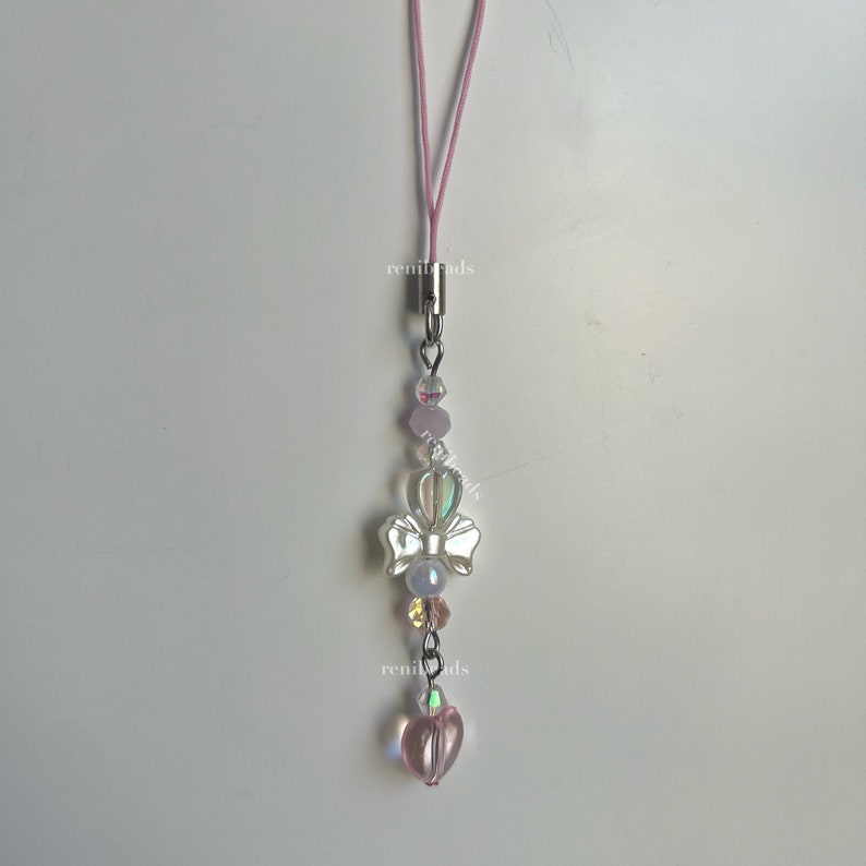 May include: A pink and white phone charm with a silver bow and heart charm. The charm is attached to a pink string.