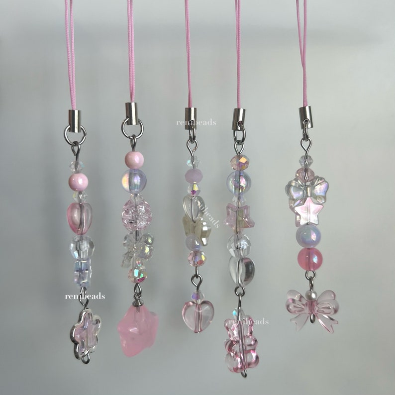 May include: Five pink and clear charm phone straps with different charms, including a star, a heart, a butterfly, and a bear. The charms are made of glass and metal.