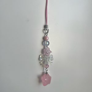 May include: A pink and white beaded phone charm with a pink star charm at the bottom. The charm features a butterfly design with iridescent beads.