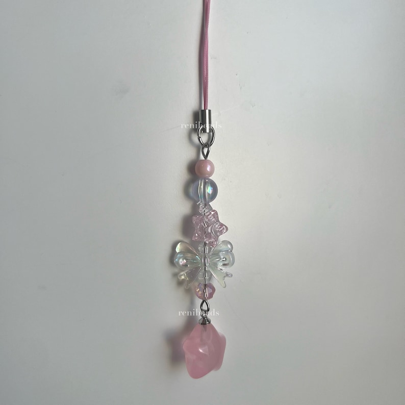 May include: A pink and white beaded phone charm with a pink star charm at the bottom. The charm features a butterfly design with iridescent beads.