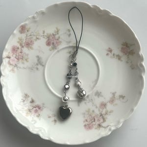 May include: A silver-toned charm with a black cord, featuring a large heart pendant and smaller heart and bead accents. The charm is displayed on a white plate with a floral pattern. The text "renibeads" is visible.