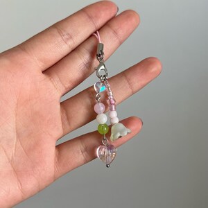 Peut inclure: Un porte-cl&eacute;s de t&eacute;l&eacute;phone en perles roses et vertes avec un charme en cristal en forme de c&oelig;ur et deux charmes en forme de fleurs blanches.