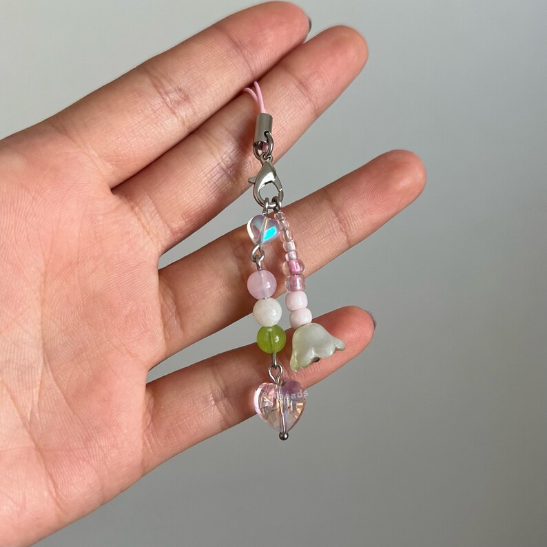 Peut inclure: Un porte-cl&eacute;s de t&eacute;l&eacute;phone en perles roses et vertes avec un charme en cristal en forme de c&oelig;ur et deux charmes en forme de fleurs blanches.