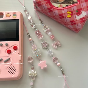 May include: Four pink and clear beaded phone charms with different charms, including a butterfly, a star, a flower, and a bow. The charms are attached to a pink string.