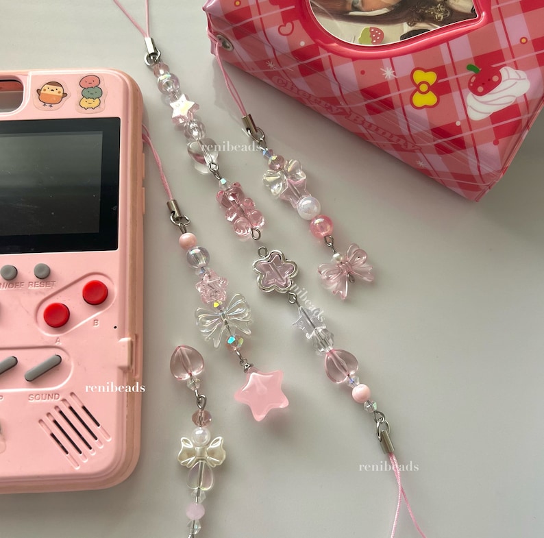 May include: Four pink and clear beaded phone charms with different charms, including a butterfly, a star, a flower, and a bow. The charms are attached to a pink string.