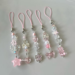 May include: Five pink and clear beaded phone charms with silver accents. Each charm features a different design, including a star, heart, flower, bow, and bear.