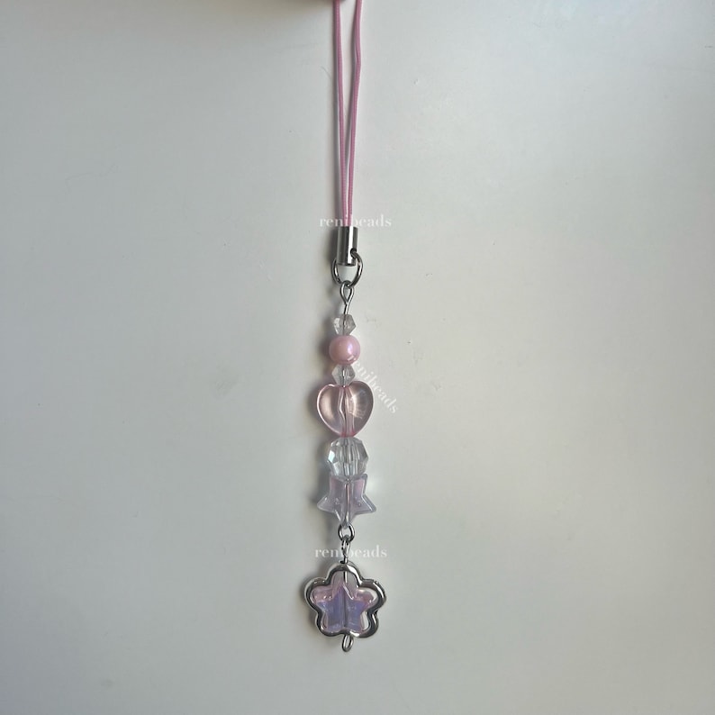 May include: A pink and clear beaded phone charm with a heart, star, and flower charm. The charm is attached to a pink string.