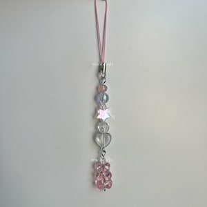May include: A pink and clear beaded phone charm with a star, hearts, and a pink gummy bear charm. The charm is attached to a pink elastic string.