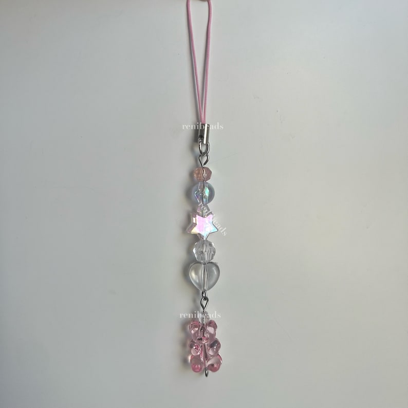 May include: A pink and clear beaded phone charm with a star, hearts, and a pink gummy bear charm. The charm is attached to a pink elastic string.