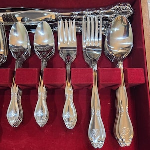May include: A set of polished silver-tone stainless steel flatware arranged in a storage case with a red velvet lining. The collection includes forks, spoons, and knives, each featuring an ornate handle design with a letter W monogram engraved.