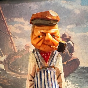May include: A hand-carved wooden figurine of a person wearing a flat cap, a red neckerchief, and blue and white striped overalls. The figure has a carved pipe in their mouth and stands on a dark brown square base, showcasing visible wood grain texture.