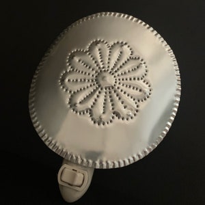 May include: A silver-colored night light with a decorative punched tin design. The night light has a circular shape with a floral pattern in the center and a scalloped edge. It features an on/off switch.
