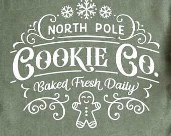 May include: An olive green sweatshirt with the text "North Pole Cookie Co. Baked Fresh Daily" in white. The design includes snowflakes, swirls, and a gingerbread man. A candy cane and a star are on the right side.