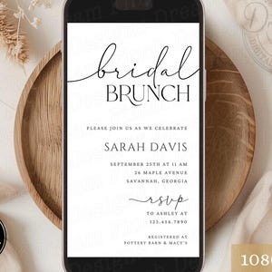 May include: A smartphone screen shows a digital invitation for a bridal brunch. The invitation is in black script with the words "bridal brunch" and event details. The phone is on a wooden tray, with a neutral background.